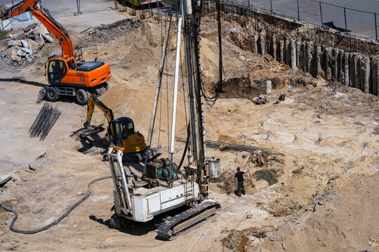 an active construction site showing a large white piling rig and an orange excavator working on deep foundation groundworks in a dirt excavation pit.