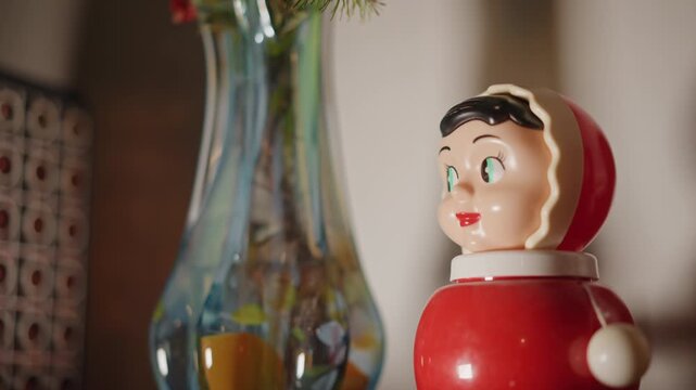 Vintage red toy beside glass vase, closeup shot of retro bobble doll on wooden shelf, warm window light, shallow depth of field, soft bokeh, glossy paint details, subtle wear and patina, cozy interior