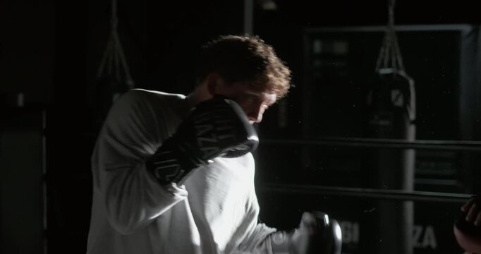 Young boxer training with a coach in a dimly lit boxing gym. Athlete practices punches, defensive movements and striking technique while wearing boxing gloves. Discipline, endurance and preparation