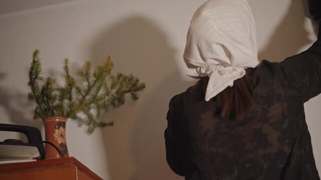 White homemaker performs evening clock ritual by wardrobe, reaching to wind and adjust cuckoo clock while iron and plant sit nearby, backview composition with warm lamp light and cozy domestic mood