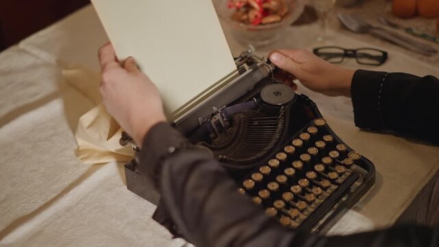 Hands loading blank paper into typewriter, cozy desk scene with linen tablecloth and glasses, author adjusting carriage and ribbon, warm amber light, closeup on mechanical keys and tactile ritual