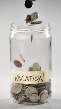 Close-up shot of coins falling into a coin jar labeled 'Vacation' in slow motion. Vertical Video.