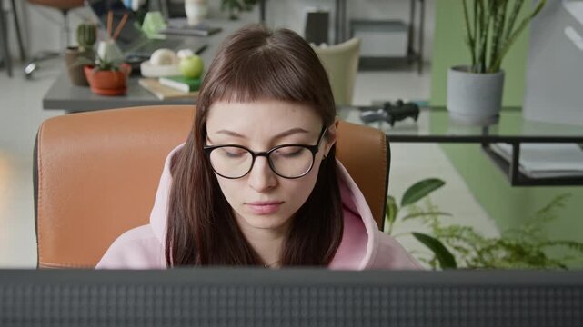 High-angle close-up of face of young gen Z Caucasian female web developer in glasses working on desktop computer in office, writing script, eyes observing screen concentrating on task, drinking coffee