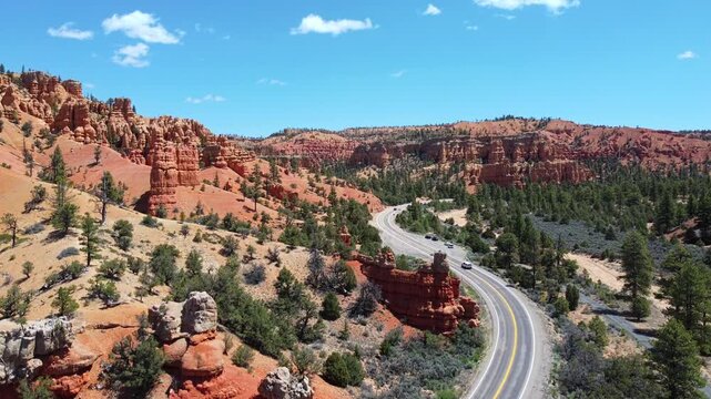 Smooth drone footage flying over a winding highway with cars amidst brilliant red rock spires and pinnacles in Dixie National Forest. Magical canyon landscape in Utah under a sunny blue sky.