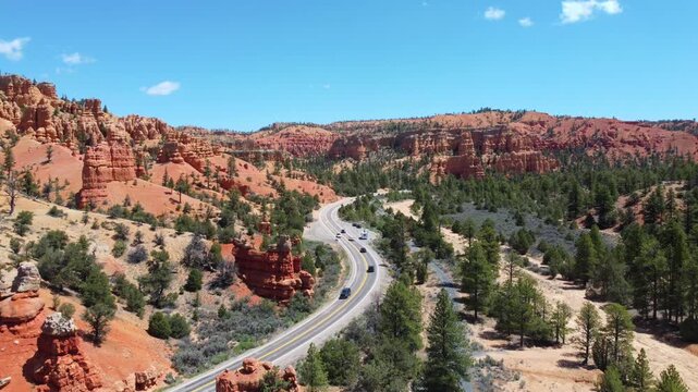 Smooth drone footage flying over a winding highway with cars amidst brilliant red rock spires and pinnacles in Dixie National Forest. Magical canyon landscape in Utah under a sunny blue sky.