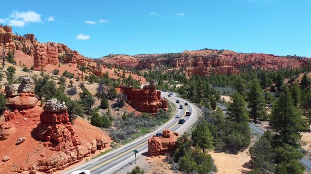 Smooth drone footage flying over a winding highway with cars amidst brilliant red rock spires and pinnacles in Dixie National Forest. Magical canyon landscape in Utah under a sunny blue sky.
