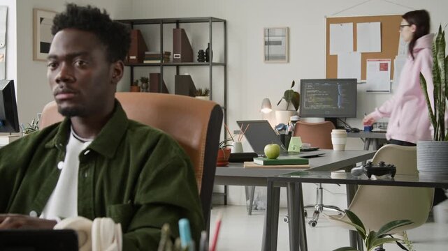 Medium rack focus of African American male programmer writing script on computer, Caucasian female colleague walking in, sitting at desk, putting on headphones while working together in office