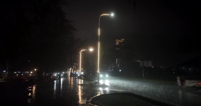 Heavy rain falls on a city street at night, where cars, motorbikes, and tuk-tuks drive under illuminated lamp posts, casting reflections on the soaked, shimmering road.
