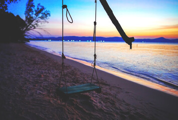 Fototapeta premium Swing on sea beach on sunset. Swing hangs on tree branch on shore tropical resort. Beach swing at ocean waves on sunset background. Vacation at sea with love on tropical paradise. 