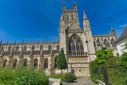 A historic stone cathedral with tall gothic windows and a prominent central tower stands bathed in sunlight under a vivid blue sky at Gloucester Cathedral - Gloucester - Gloucestershire - UK