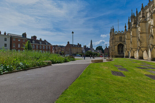 A grand Gothic cathedral stands beside a paved path and grassy area. Beyond, historic town buildings line a street at Gloucester Cathedral - Gloucester - Gloucestershire - UK