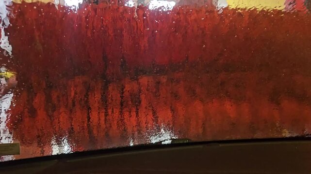 View from inside a car during an automatic car wash, showing red rotating brushes, soap foam and water droplets streaking across the windshield for a dramatic abstract texture and motion effect.