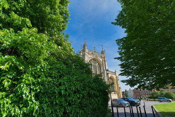 Naklejka premium A historic stone cathedral with gothic architecture is framed by lush green trees against a bright blue sky at Gloucester Cathedral - Gloucester - Gloucestershire - UK