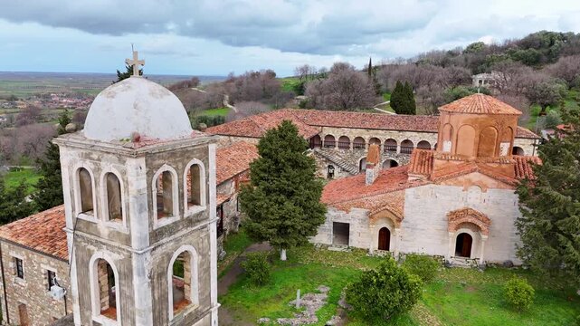High-angle aerial establishing shot of the historic concrete bell tower, Byzantine brick domes, and stone cloister courtyard of St. Mary Church at Apollonia Archaeological Park, Albania