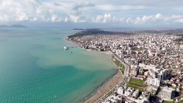 Wide aerial tracking shot flying forward over dense coastal high-rises, sandy beaches, and the turquoise waters of the Bay of Vlor&euml; under a cloudy blue sky in Albania