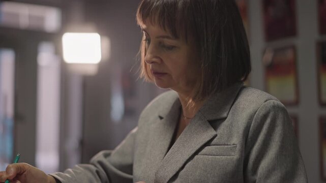 Female caucasian composer editing score under studio lights, closeup hands with pen on manuscript, thoughtful focused expression, grey blazer and illuminated poster backdrop, quiet creative editing