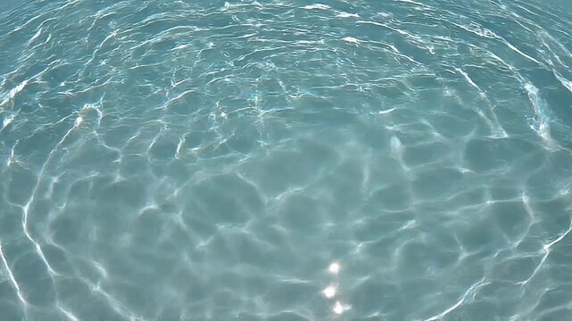 Close-up overhead view of clear turquoise water surface with concentric ripples and light reflections creating a abstract pattern