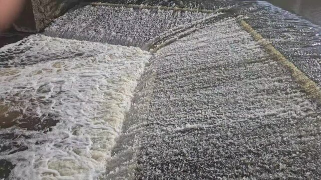 Aerial perspective of water flowing over a concrete weir, revealing a sharp contrast between foamy, turbulent spillway and calm, reflective downstream surface with visible ripples and texture.