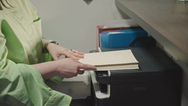 Music studio copy station preparing scores with hands feeding binder into printer and flipping pages to check notation, assistant in green uniform inspects document, compact machine and tray visible,