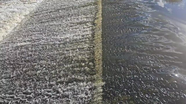 Aerial perspective of water flowing over a concrete weir, revealing a sharp contrast between foamy, turbulent spillway and calm, reflective downstream surface with visible ripples and texture.