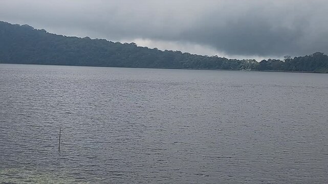 Calm tropical lake with misty rainforest hills and dramatic overcast clouds in Bedugul, Bali, Indonesia. Peaceful freshwater landscape with natural scenery and tranquil atmosphere.