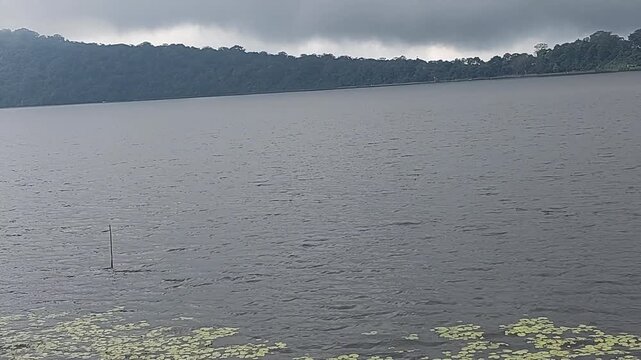 Calm tropical lake with misty rainforest hills and dramatic overcast clouds in Bedugul, Bali, Indonesia. Peaceful freshwater landscape with natural scenery and tranquil atmosphere.