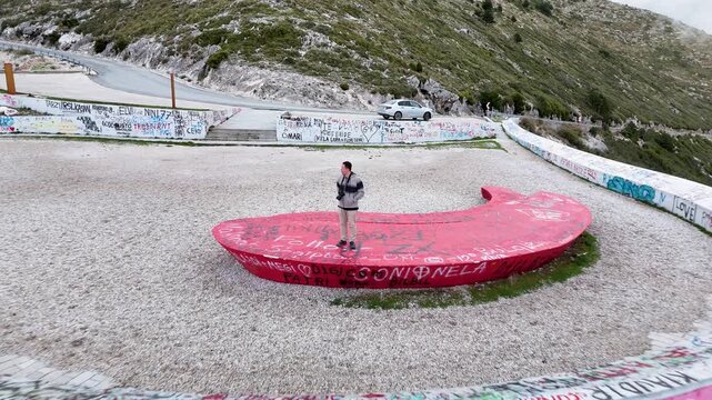 Aerial tracking shot pulling back and rising over host Charles Huang of DIY Destinations as he stands on a red graffiti-covered crescent platform at the mountain pass viewpoint in Albania
