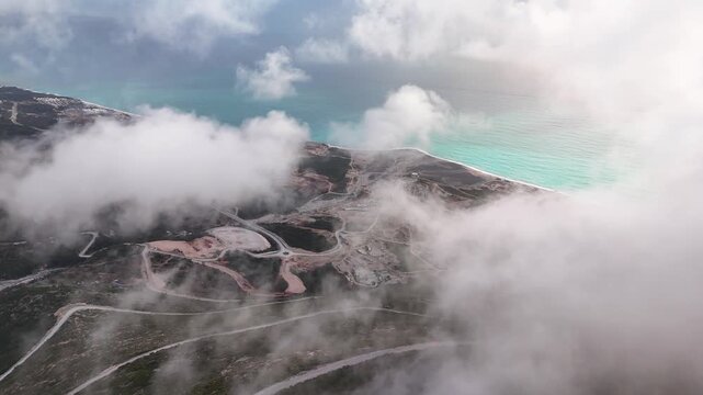 High-angle aerial shot moving forward through low clouds over a rugged mountain highway, hillside construction zones, and the turquoise Ionian Sea at Llogara Pass, Albania