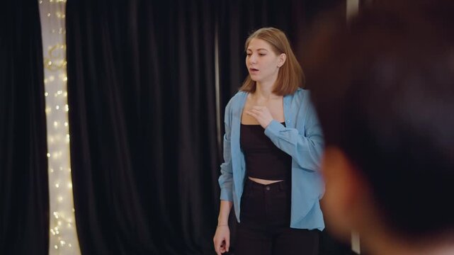 Caucasian female actor auditioning onstage with black curtain and fairy lights, thoughtful expression and hand gesture, blurred audience foreground, intimate school theatre atmosphere