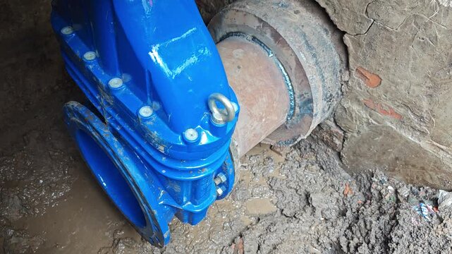 Close-up of a heavy-duty blue industrial gate valve connected to a water pipeline in a muddy trench. Utility infrastructure repair and maintenance