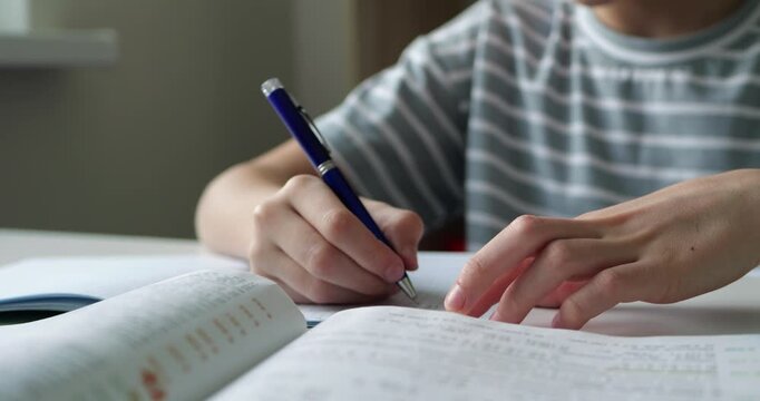 Close-up of student hands writing notes in notebook with textbook on desk. Horizontal shot capturing focused study session, education and learning environment at home or school.