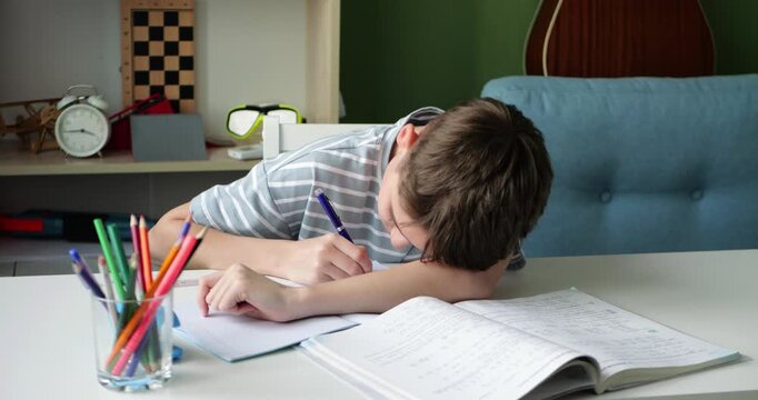 Tired school boy reluctantly doing homework at home while writing in notebook with bored expression. Young student resting head on hand during study time in cozy room. Child does not want do homework