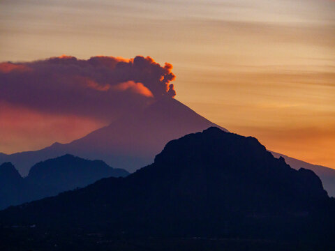 Silueta del Cerro de Tepoztl&aacute;n en primer plano y el volc&aacute;n Popocat&eacute;petl activo de fondo con fumarola al atardecer