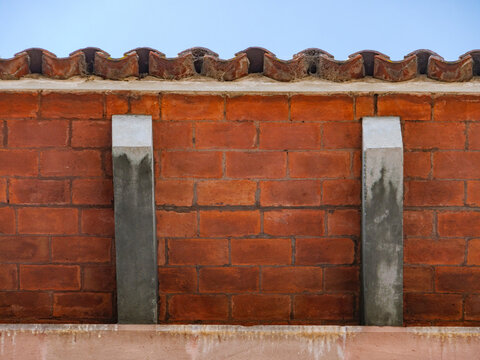 Muro r&uacute;stico de ladrillo rojo con pilares de concreto y tejas de barro bajo un cielo azul, detalle arquitect&oacute;nico.