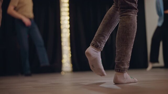 Closeup barefoot footwork across wooden floor during dance workshop, highlighting toe placement, pivoting, balance shifts, and cadence as group rehearses under warm stage lighting.