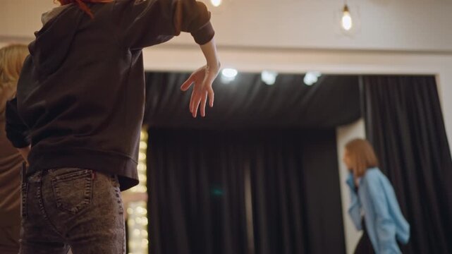 White redhead dancer practicing choreography in studio, choreographer leads focused session with hoodie and jeans attire, close camera on hand cues and timing, string lights framing black curtain,