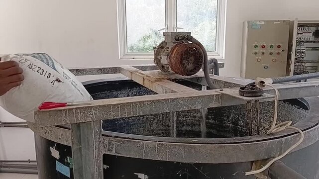 A worker's hands pouring chemical powder from a white bag into a large industrial mixing tank with a running agitator for water treatment.
