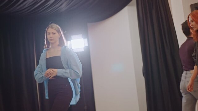 Female caucasian student walks into audition corridor toward studio lights, lineup with redhead coach and peers, wardrobe layers, nervous anticipation, casting rehearsal with director observing