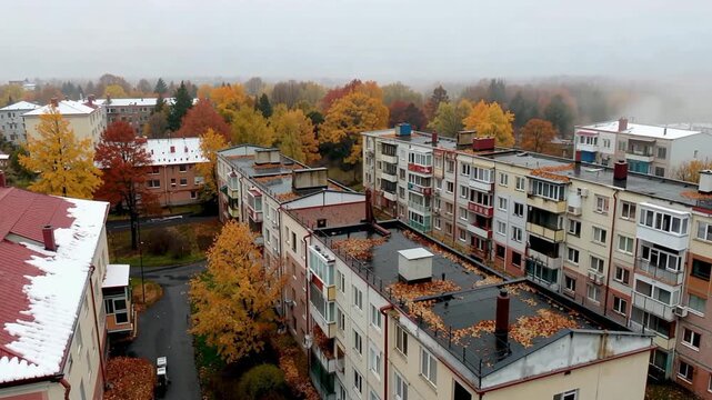 Drone captures urban area filled with residential buildings surrounded by vibrant autumn foliage. Mist lingers in the air creating an atmospheric setting