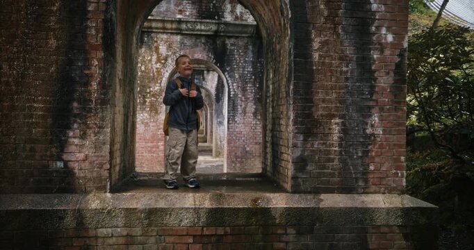 Dolly away from little boy with backpack standing under arch beneath bridge