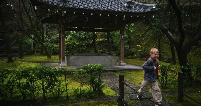 Little boy leaving beautiful, old Japanese structure in lush forest area - wide shot