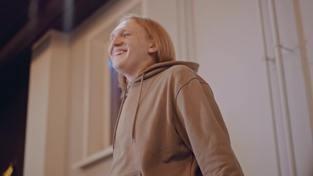 Songwriter pausing by window in studio, hoodieclad musician smiles during reflective moment, soft golden light and high ceiling studio set inspire lyrical ideas, quiet breakthrough and creative