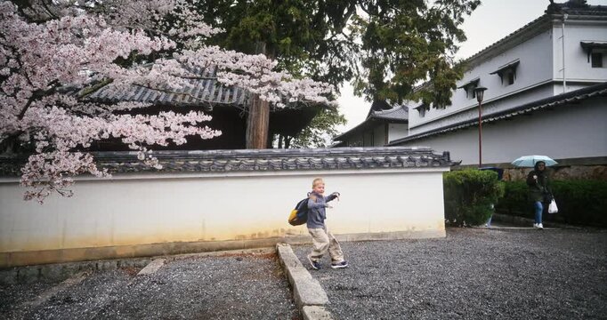 Little boy with backpack running up steps in old Japanese village near spring cherry blossoms - steady cam tracking shot