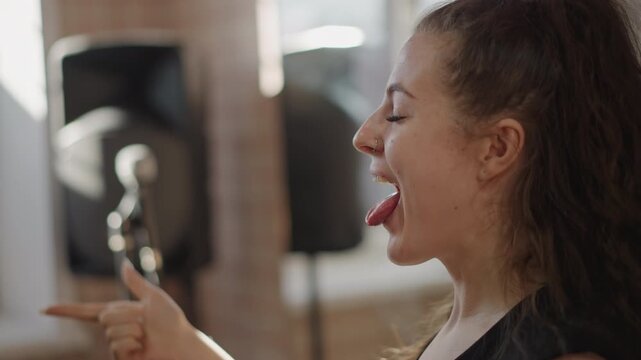 Playful singer practicing tongue articulation and expressive phrasing in bright rehearsal studio, smiling student points and sticks out tongue during energetic warmup, speaker and mic stand visible,