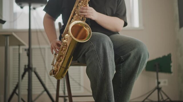 White musician leg and saxophone closeup during technique demonstration, teacher focusing on fingering and posture on wooden stool, textured carpet and amp visible, calm instructional vibe