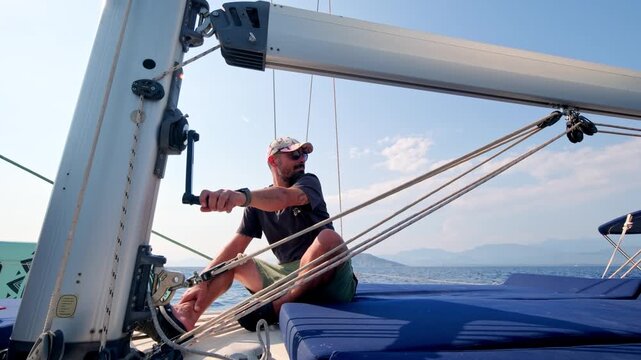 Man sitting on the deck of a sailing yacht while handling rigging equipment during a sea voyage. Nautical activity, sailing lifestyle, yacht operation and summer boating adventure.