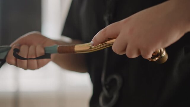 Young musician prepping saxophone with cloth and strap closeup, hands carefully cleaning mouthpiece and polishing brass, classroom light, instructor guidance visible in background, focused rehearsal