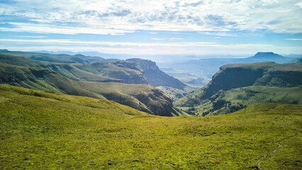Obraz premium Sunlit Drakensberg Valley Opens Toward Horizon, Broad Meadow Slopes And Layered Plateaus, Soft Cloud Cover