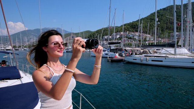 Young woman filming sailboats with a digital camera at a sunny marina. Travel content, summer vacation, yacht harbor, sailing lifestyle and relaxed seaside leisure.