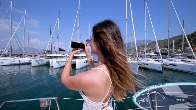Young woman filming sailboats with a digital camera at a sunny marina. Travel content, summer vacation, yacht harbor, sailing lifestyle and relaxed seaside leisure.